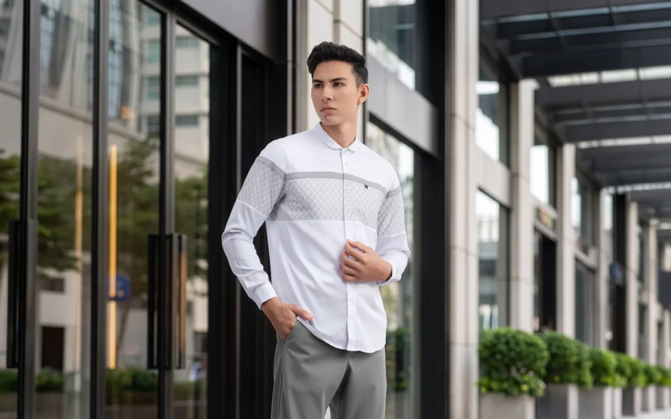 Model pria mengenakan baju kemeja putih Fadkhera tipe koko modern Oman dan celana pleated trousers abu-abu untuk acara semi-formal.